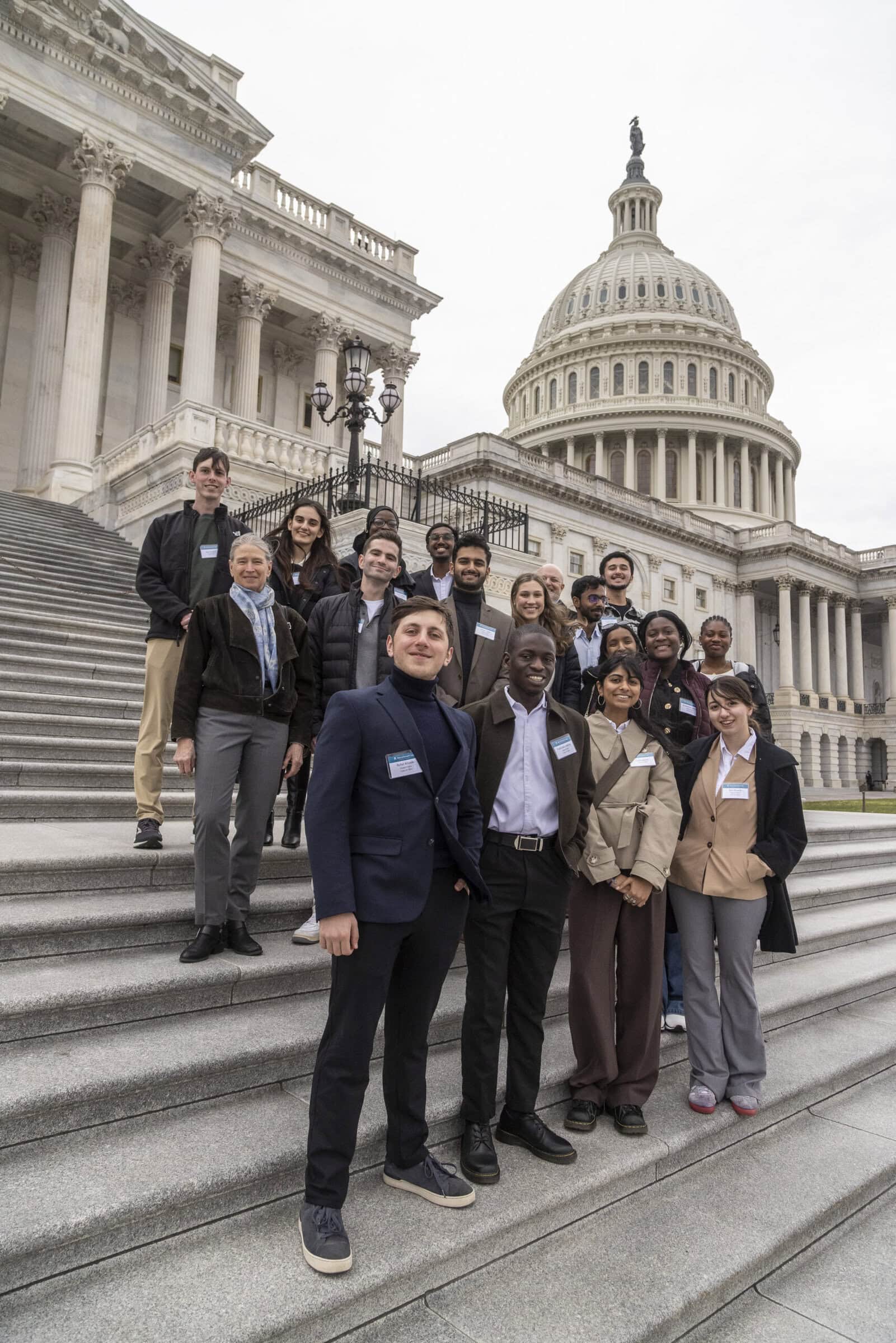 A group of about a dozen students in business attire stand and smile on the steps of the U.S. Capitol on an overcast day.