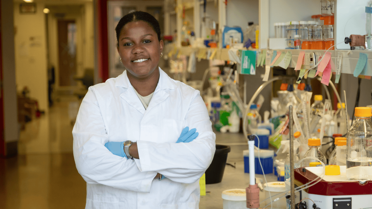 Standing in the lab, Tarynn Neal ’26 smiles with her arms crossed. She is wearing a lab coat and gloves.