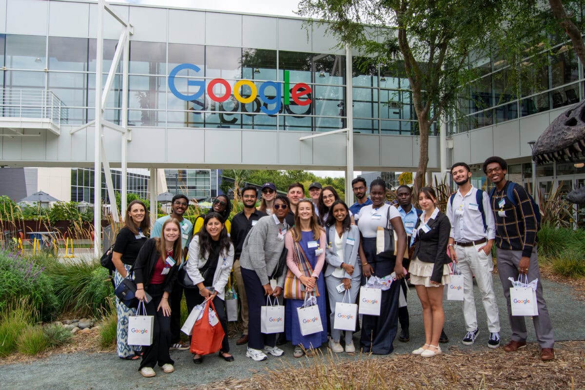 A group of Global Fellows and Ousley Naseman stand together outdoors in front of a Google sign, surrounded by trees with autumn foliage.