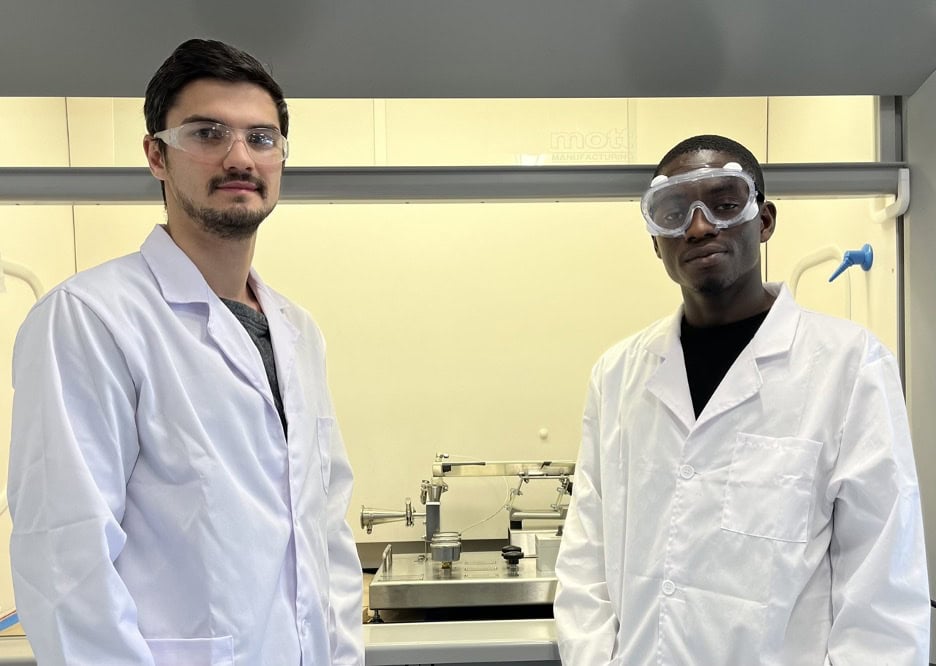 Two students in lab coats stand in front of lab equipment.