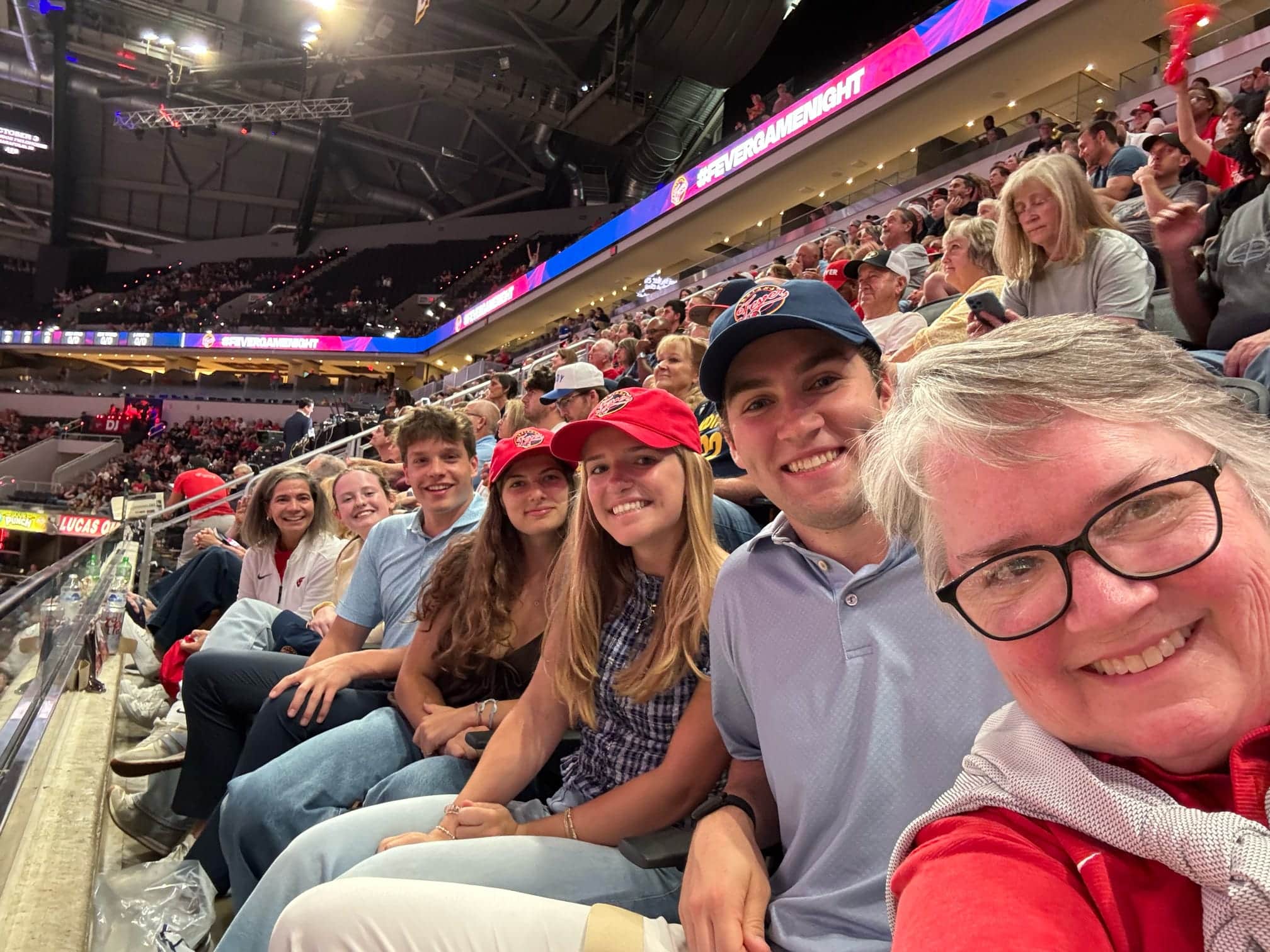 Members of the Civic Collaboration team sit together in the stands at an Indiana Fever game in Indianapolis, smiling for a selfie as fans fill the arena behind them.