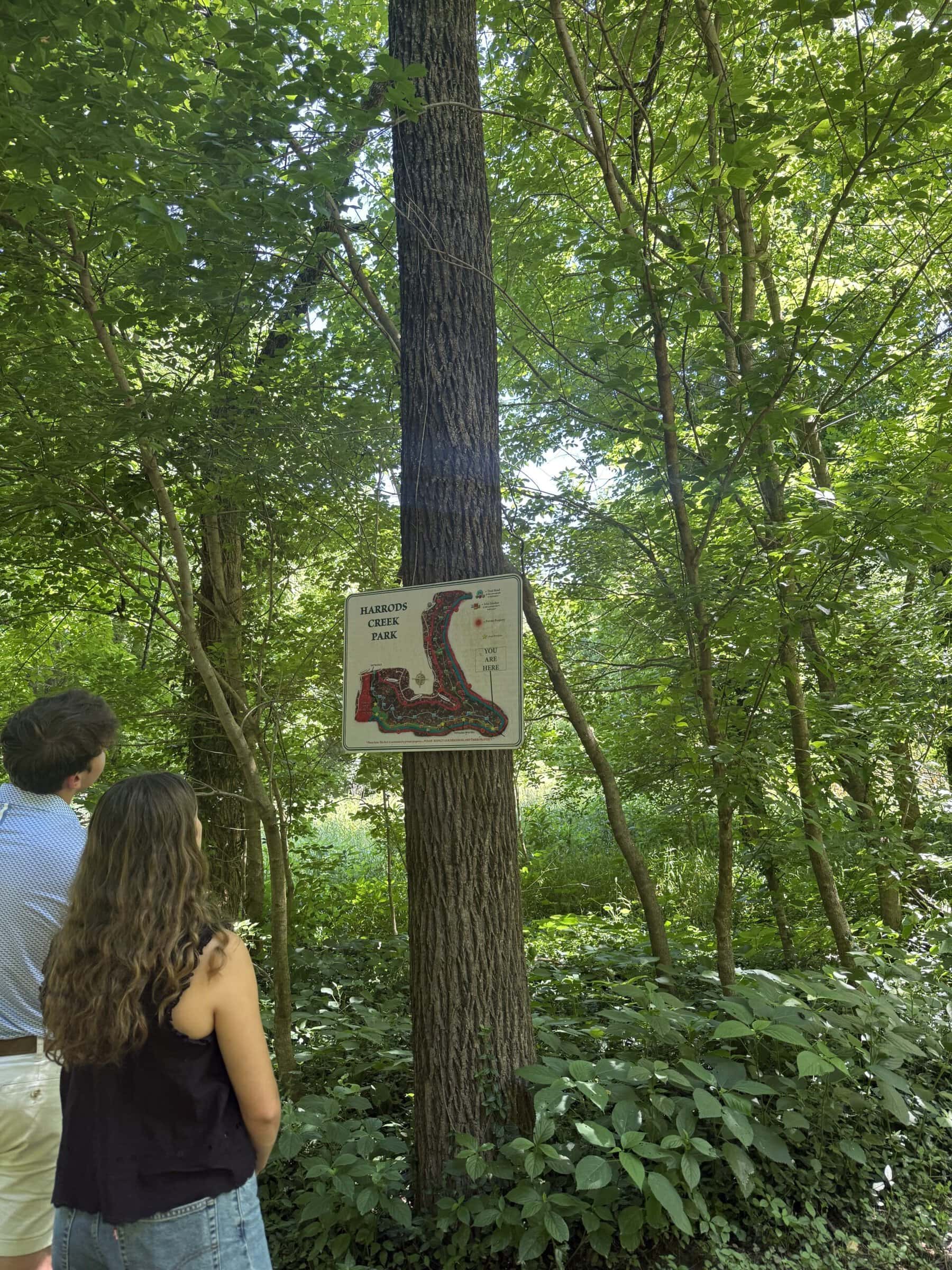 Two Civic Collaboration team members stand on a wooded trail near a sign for Harrods Creek Park, looking into the forest.