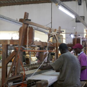 Two textile workers operate a large wooden loom inside a bright atelier filled with weaving equipment and fabric.