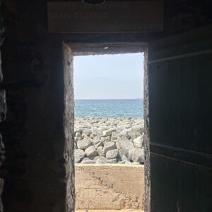 A doorway marked “Door of No Return” opens from the dark interior of the Maison des Esclaves toward the Atlantic Ocean and rocky shoreline on Gorée Island, Senegal.