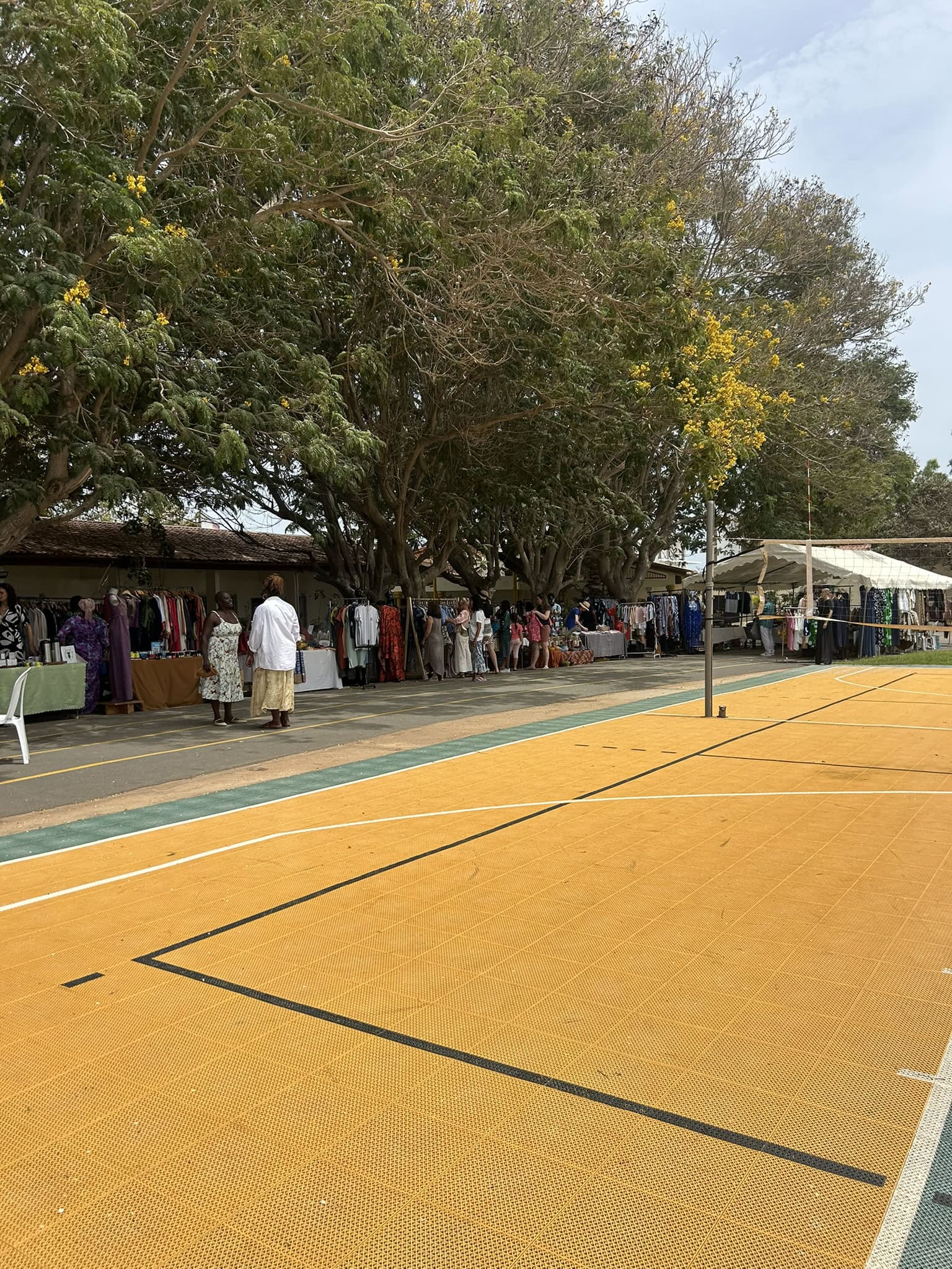 Clothing vendors display garments beneath trees beside an outdoor basketball court at an outdoor market.