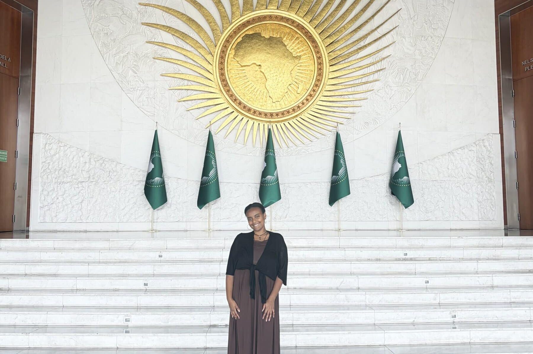 Elizabeth Alene ’28 stands beneath a large gold African Union emblem and in front of African Union flags at the organization’s headquarters in Addis Ababa, Ethiopia.