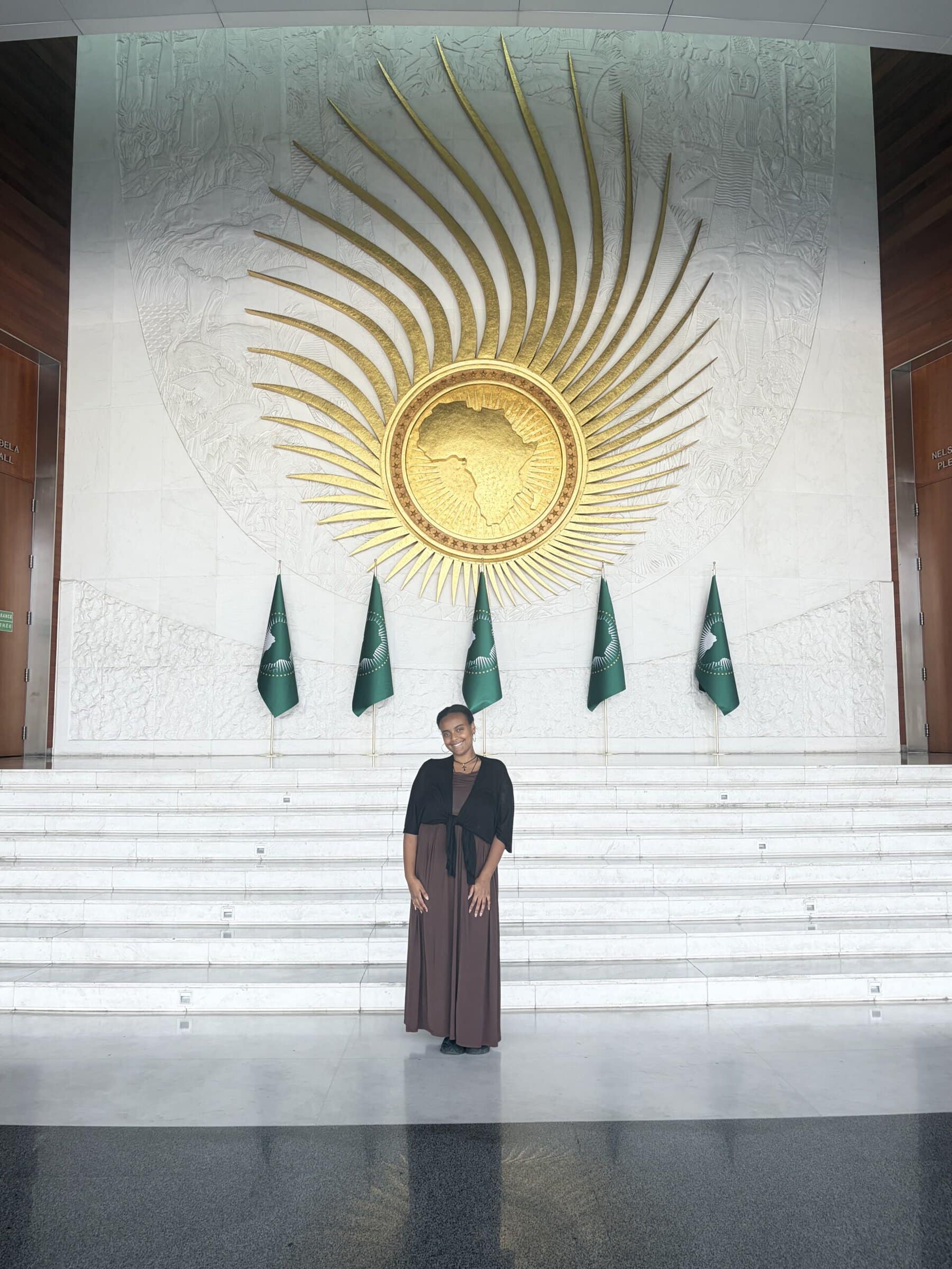 Elizabeth Alene ’28 stands beneath a large gold African Union emblem and in front of African Union flags at the organization’s headquarters in Addis Ababa, Ethiopia.