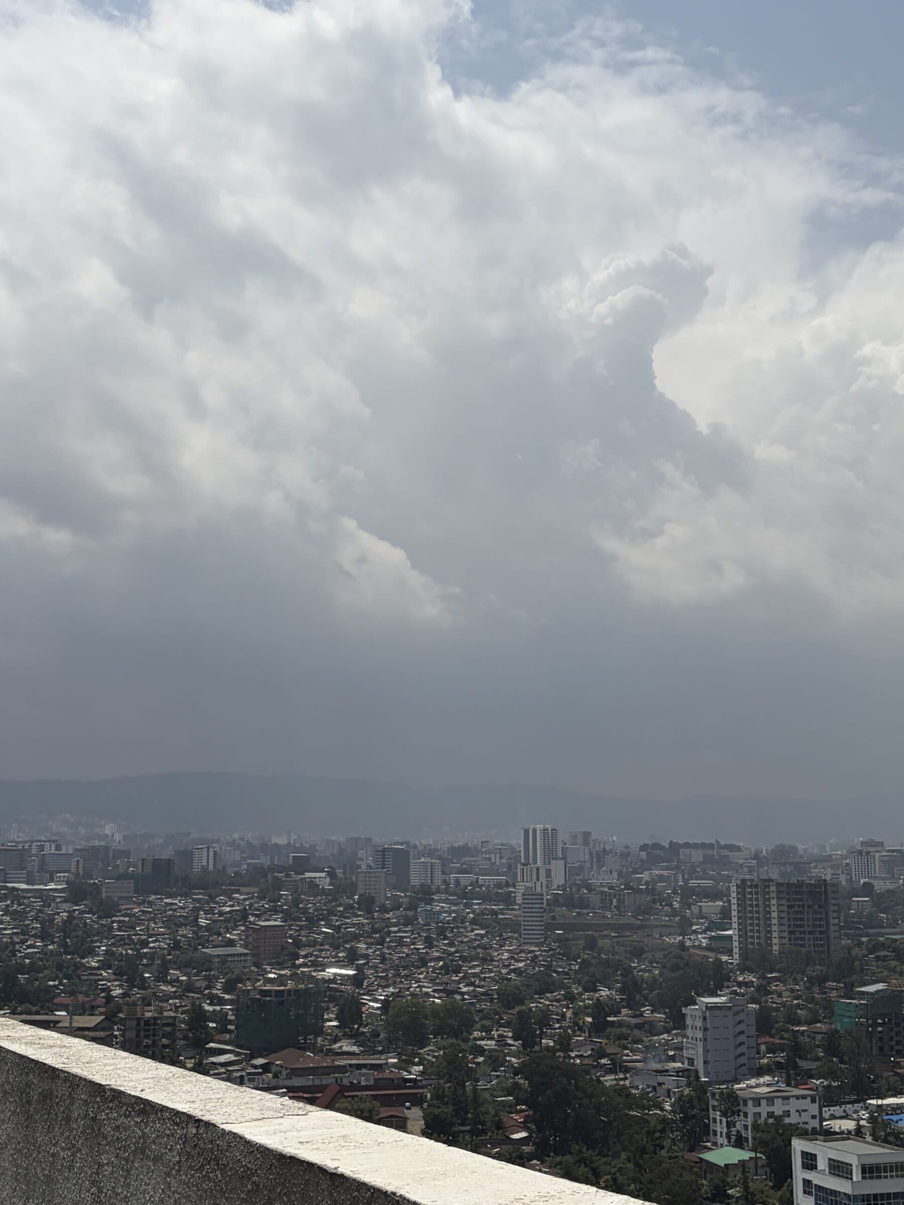 Addis Ababa stretches across a valley beneath heavy clouds, with neighborhoods, high-rise buildings and mountains visible in the distance.