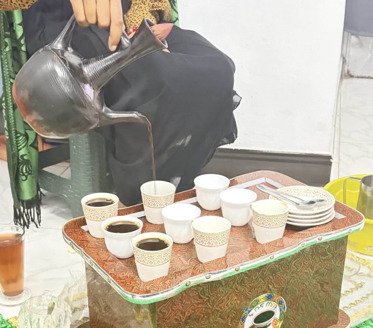 Coffee is poured from a metal pot into small cups arranged on a decorated serving tray during an Ethiopian coffee ceremony.