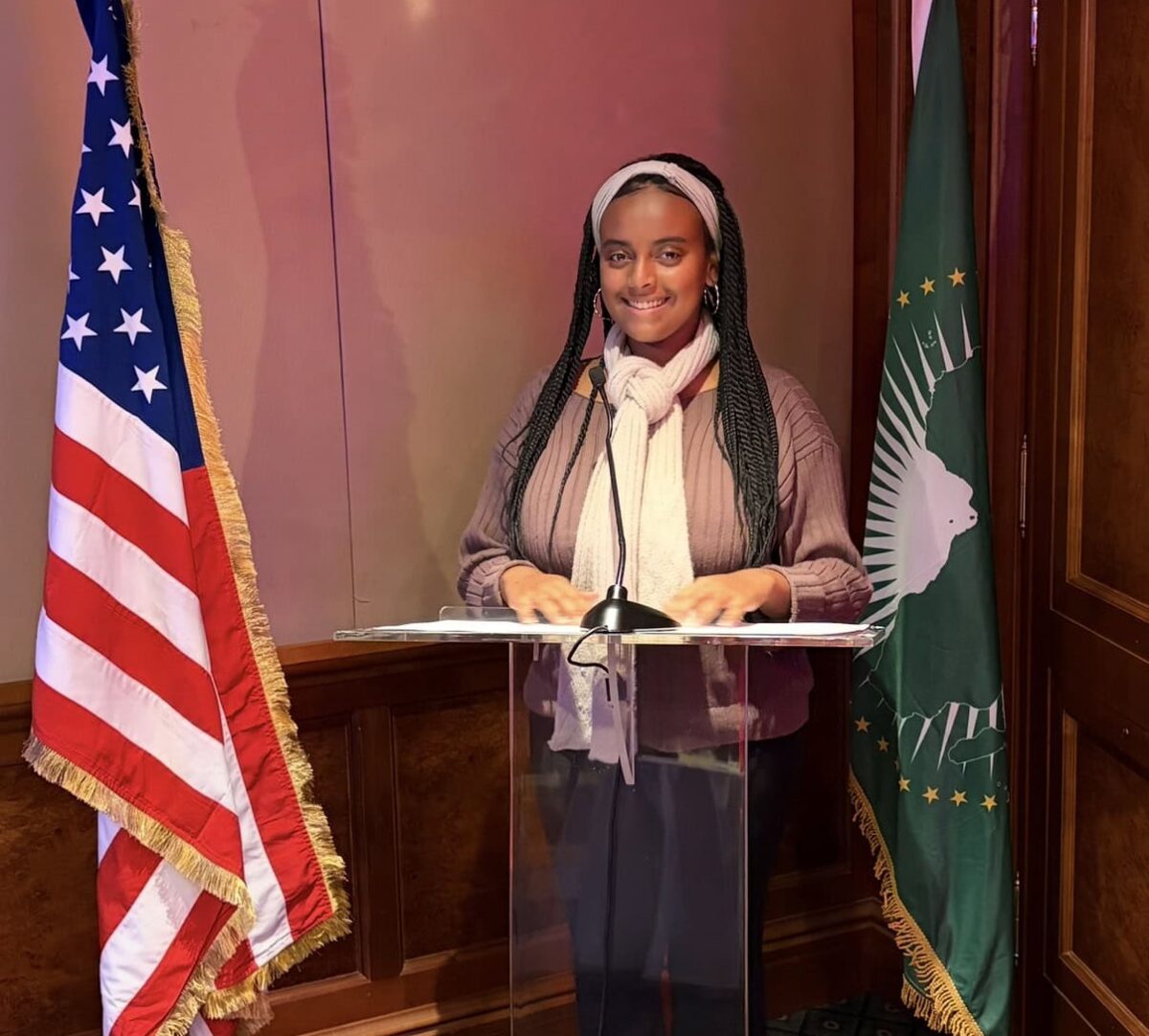 Elizabeth Alene ’28 stands at a clear lectern between U.S. and African Union flags in a wood-paneled room.