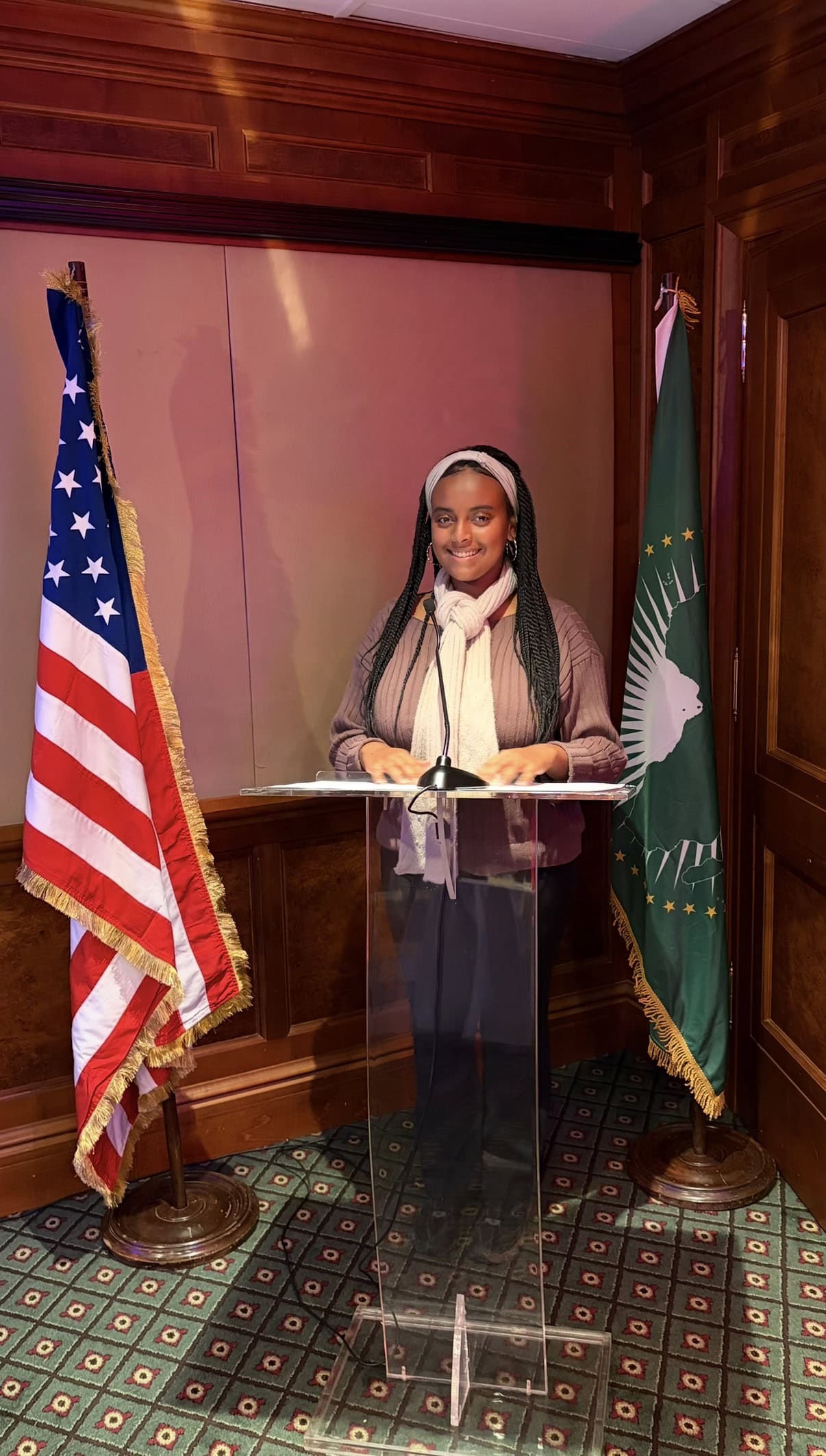 Elizabeth Alene ’28 stands at a clear lectern between U.S. and African Union flags in a wood-paneled room.