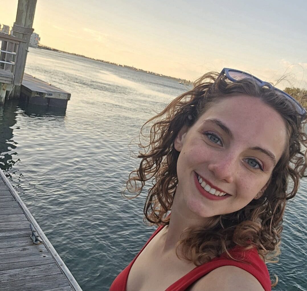 Alexandra stands on a boardwalk in Morehead City, North Carolina, with the ocean behind her as the sun sets.