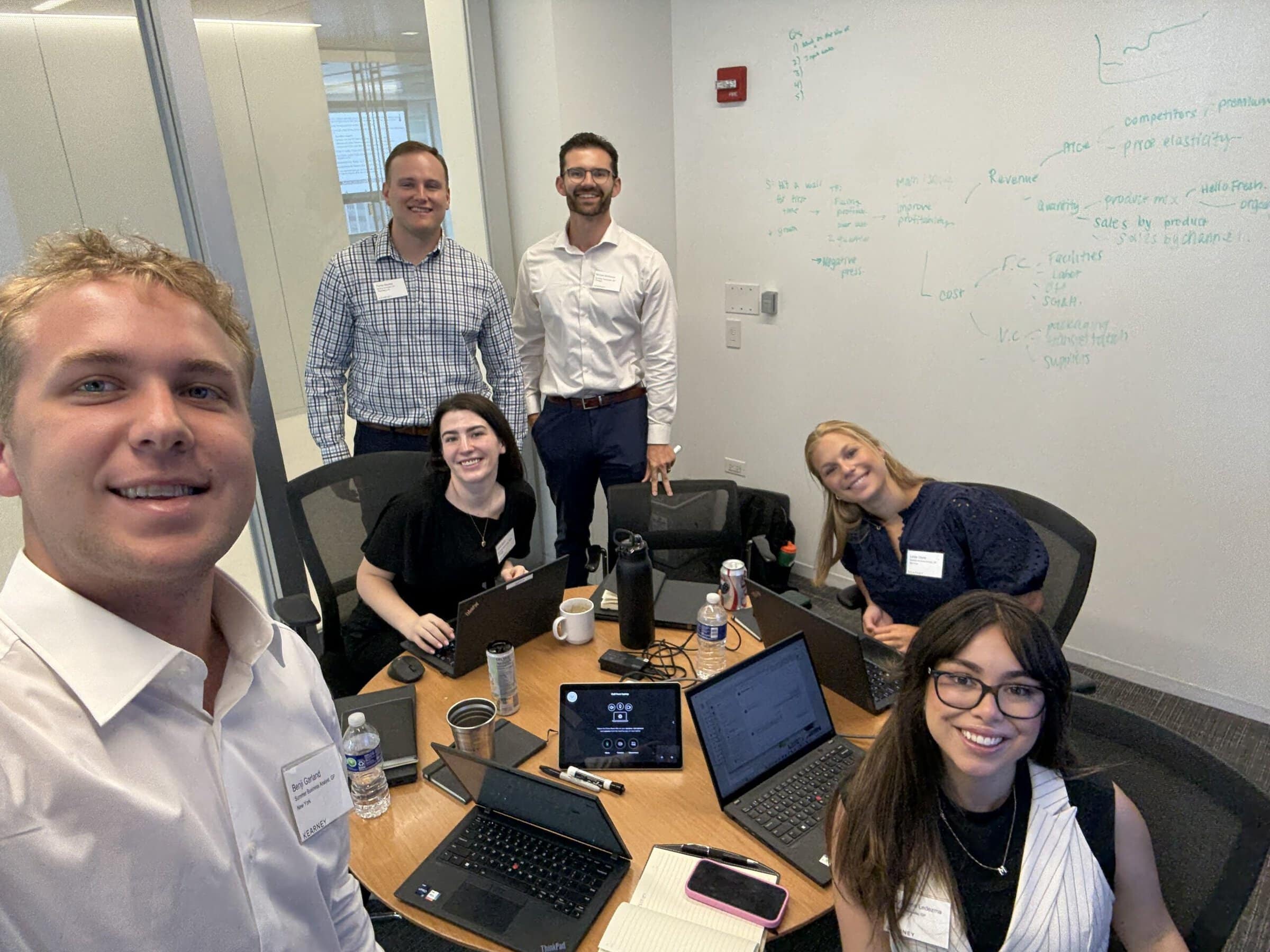 Benji Garland ’27 and five fellow interns work around a conference table during training week in Chicago, with laptops open and notes written on a whiteboard behind them.