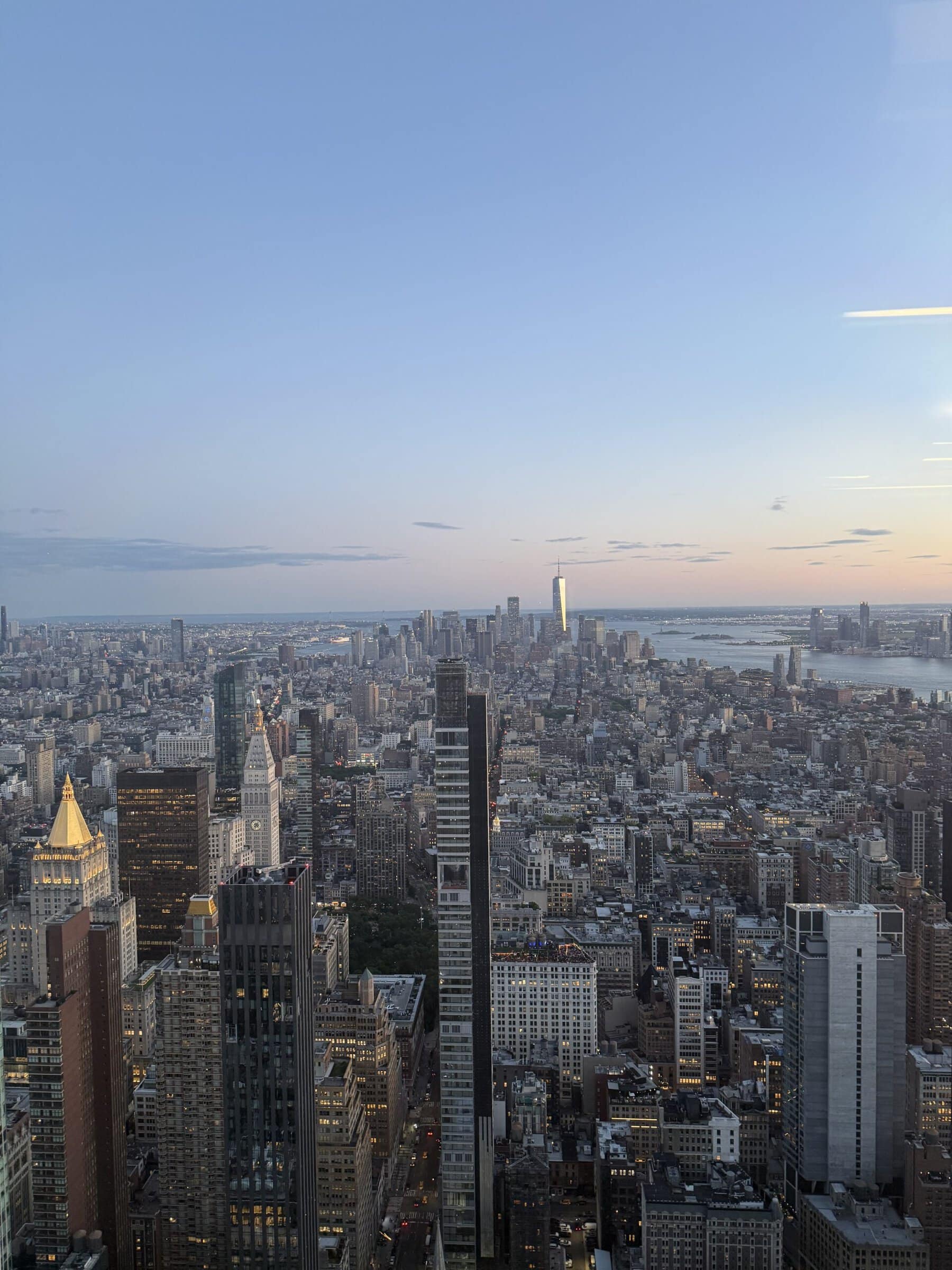 A panoramic view of New York City at dusk from the Empire State Building, with Lower Manhattan, the Hudson River and the East River visible in the distance.