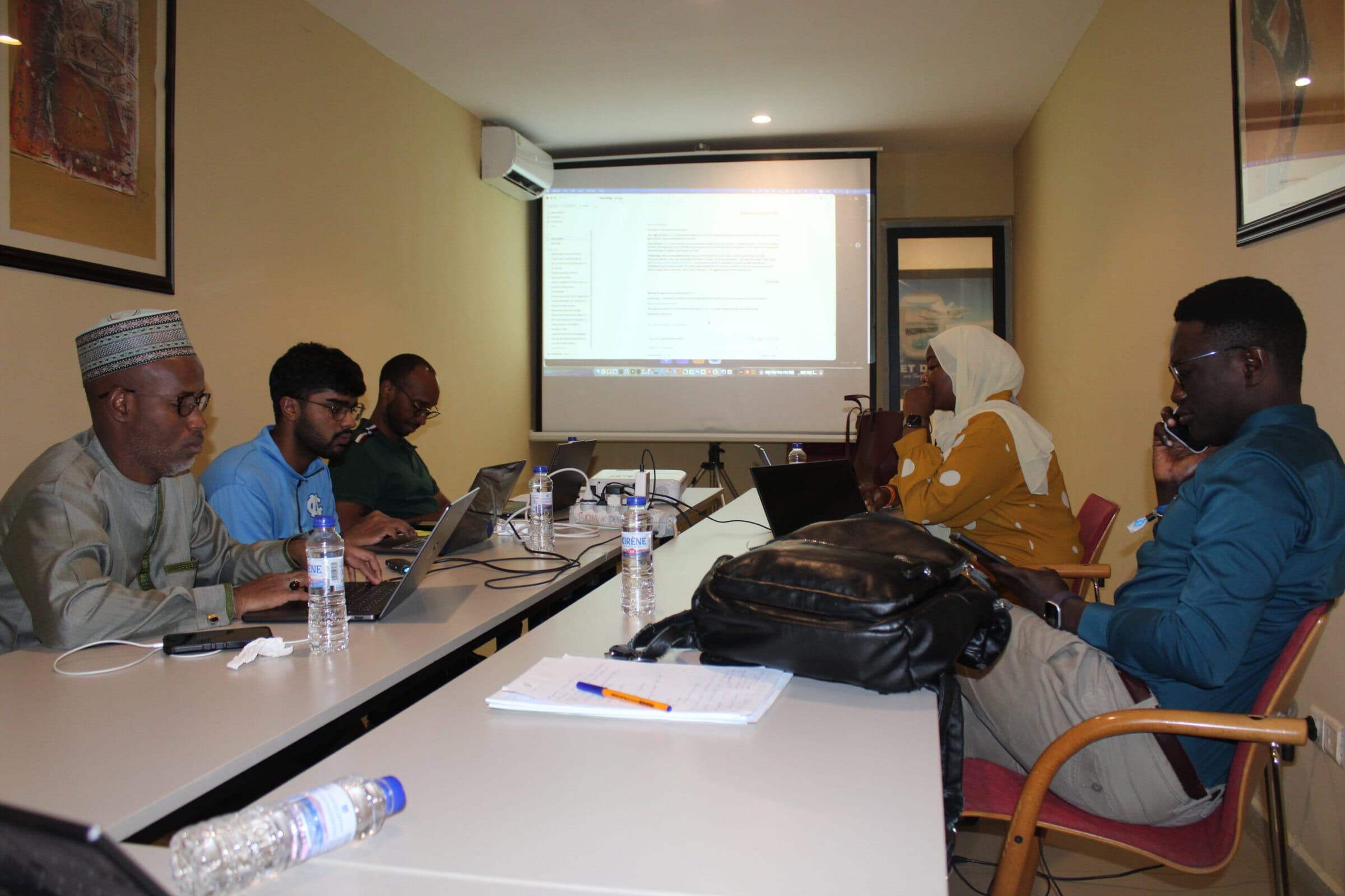 Suhan Asaigoli ’27 works with four members of the CHAI Senegal and Global Digital Health team on the Pisa app in a conference room in Saly, Senegal. Five people work on laptops as a presentation appears on the screen behind them.