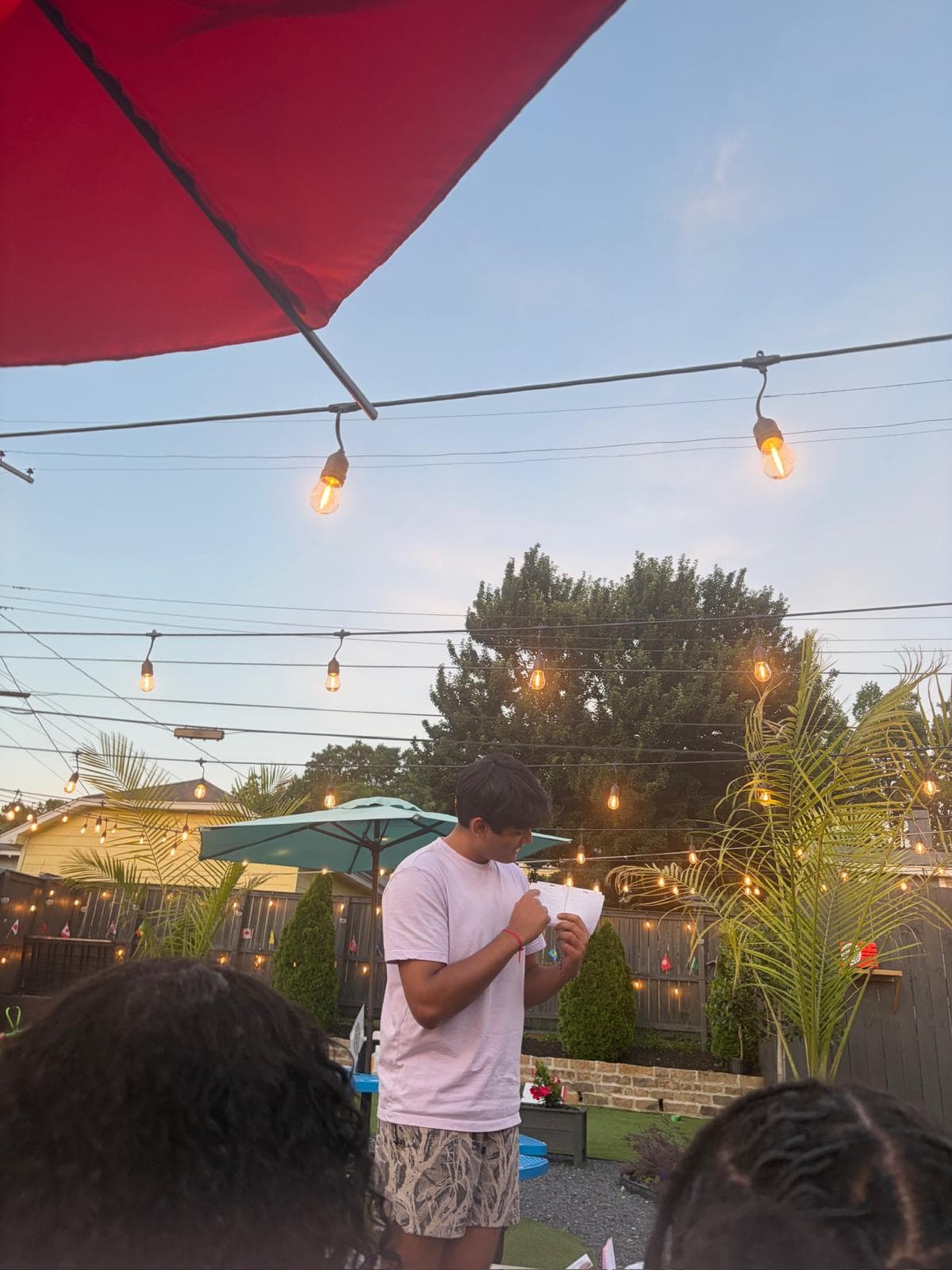 A young man in a lavender T-shirt reads from a small booklet in a backyard strung with lights and palm plants as an audience looks on.