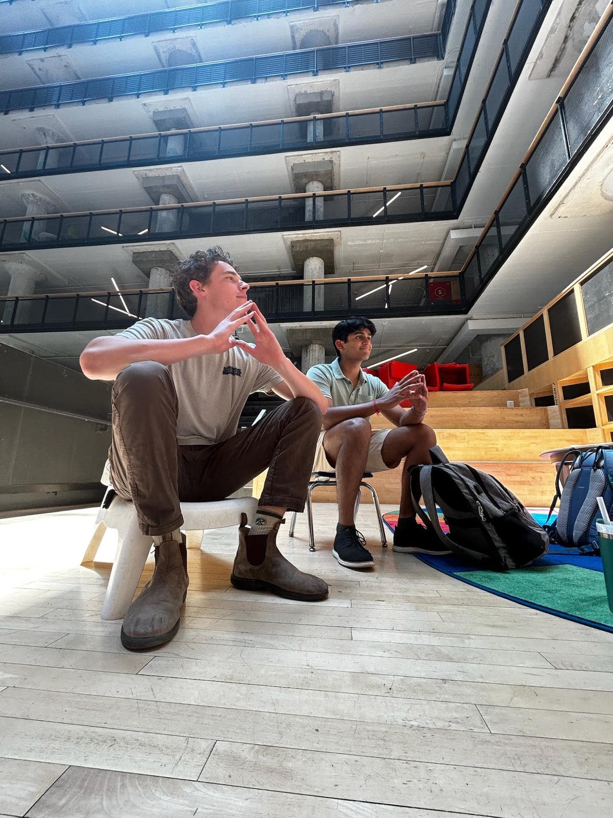 Two young men sit on low chairs in a tall atrium with open balconies rising above them, listening to someone out of frame.