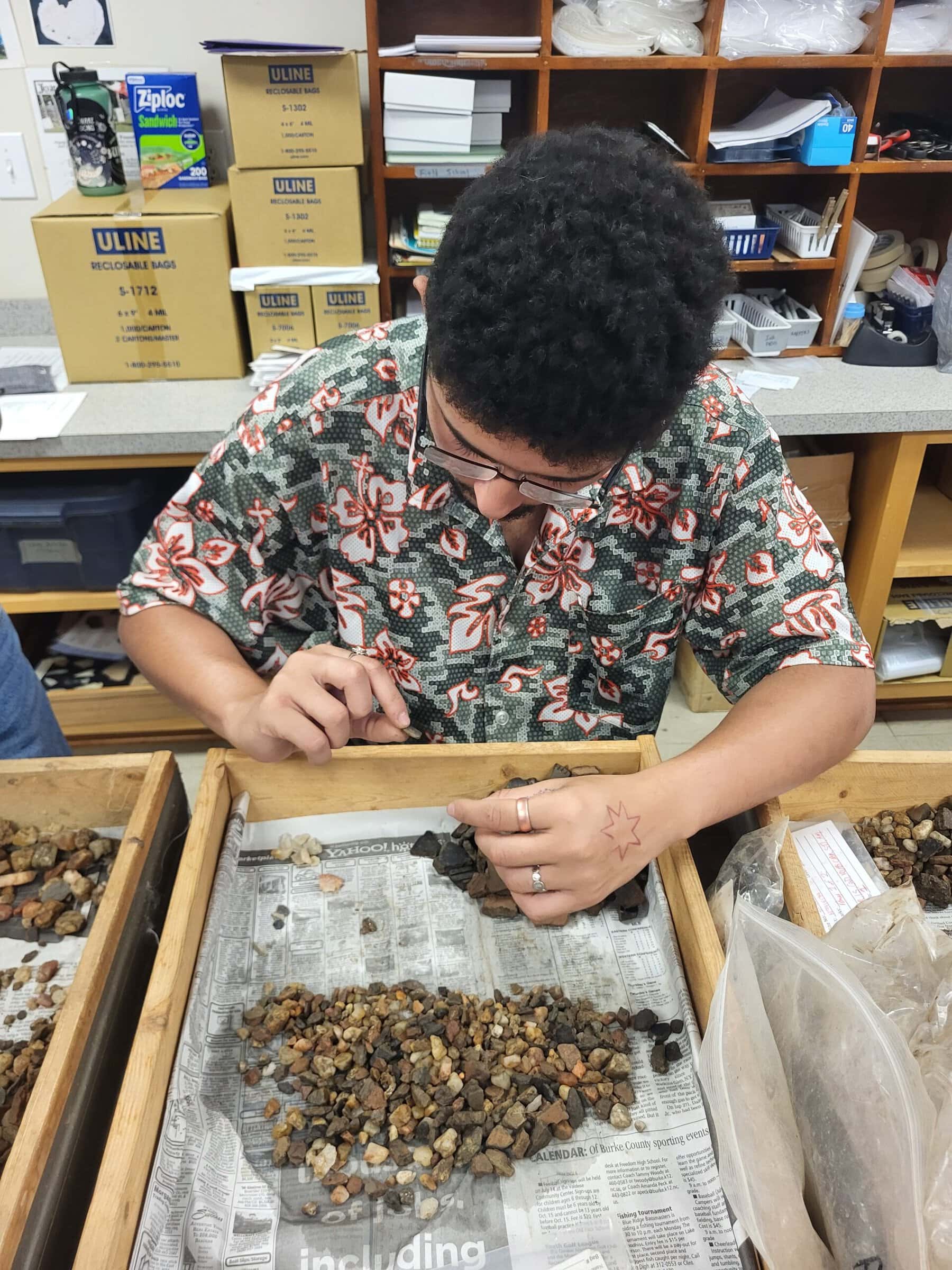 A young man in glasses and a floral shirt sorts small stones and pottery fragments in a wooden tray lined with newspaper.