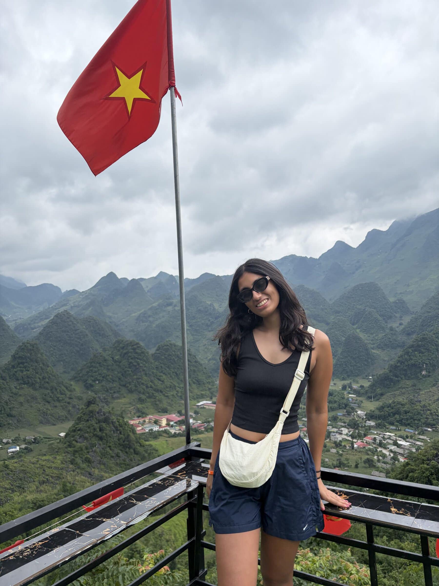 A young woman in sunglasses stands at a railed overlook beneath a Vietnamese flag, with steep green mountains and a village in the valley below.