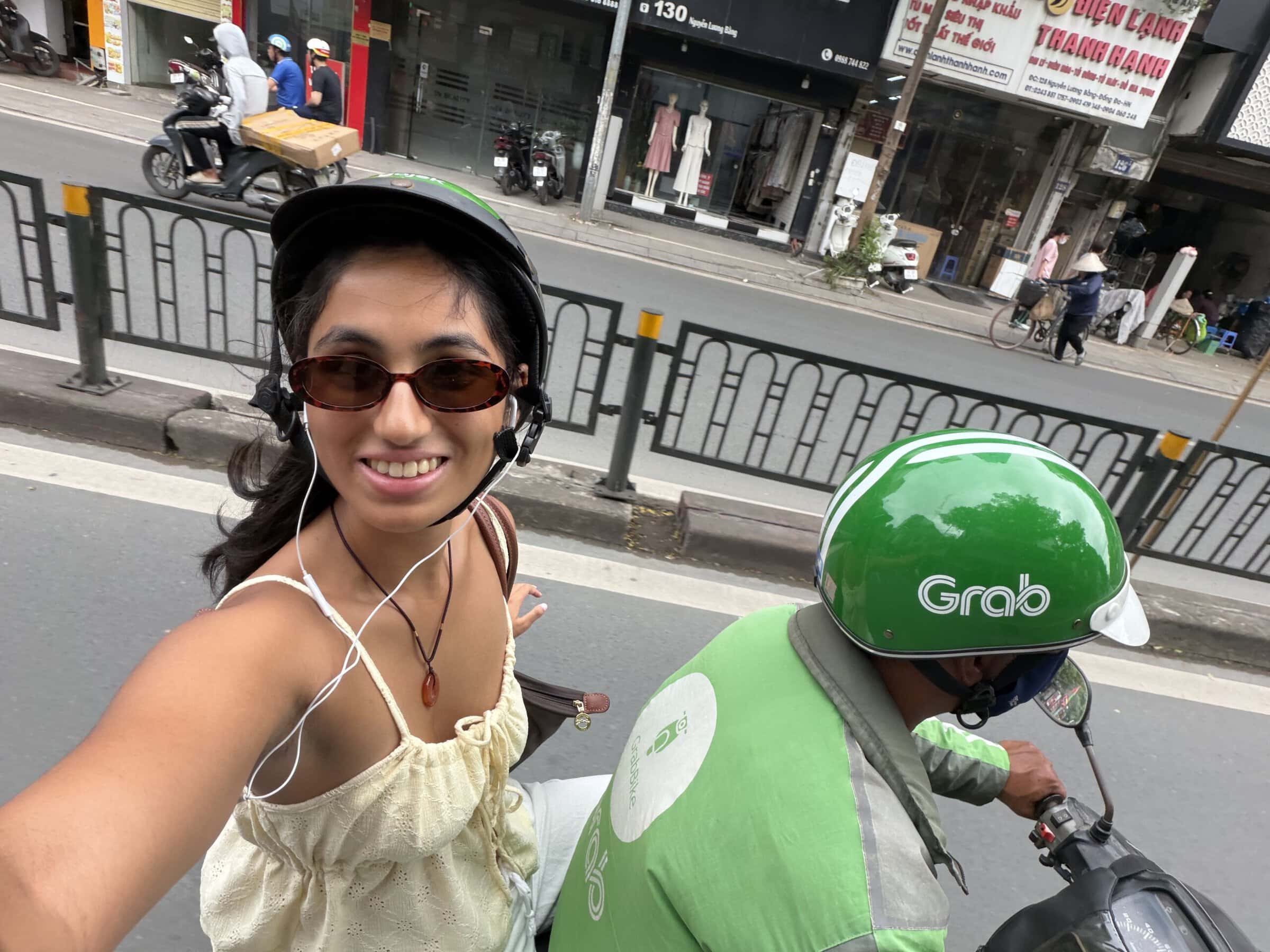 A smiling young woman in a helmet and sunglasses takes a selfie on the back of a motorbike behind a driver in a green Grab jacket and helmet on a busy city street.