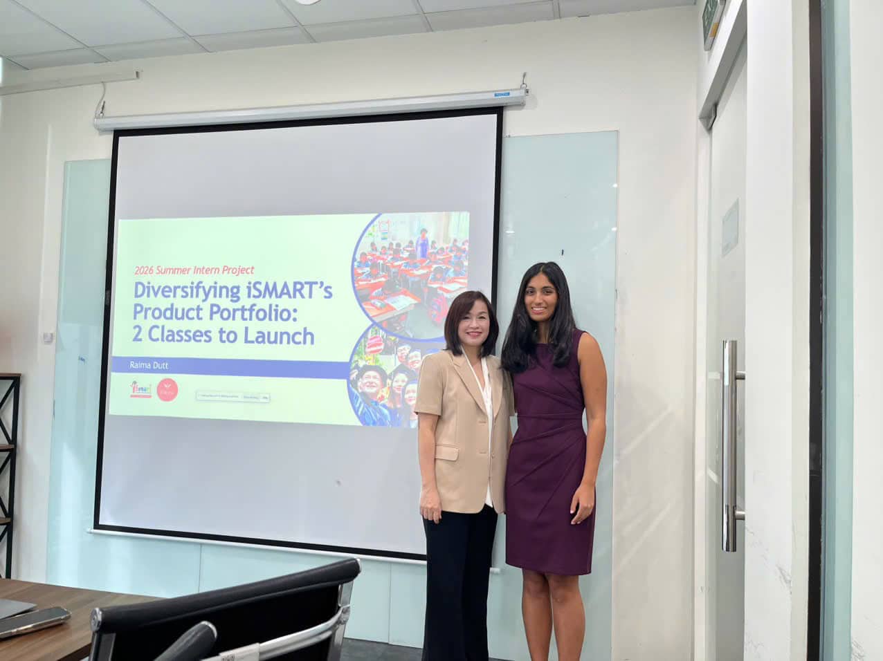 Two women stand in a conference room in front of a projected slide titled “Diversifying iSMART’s Product Portfolio: 2 Classes to Launch.”