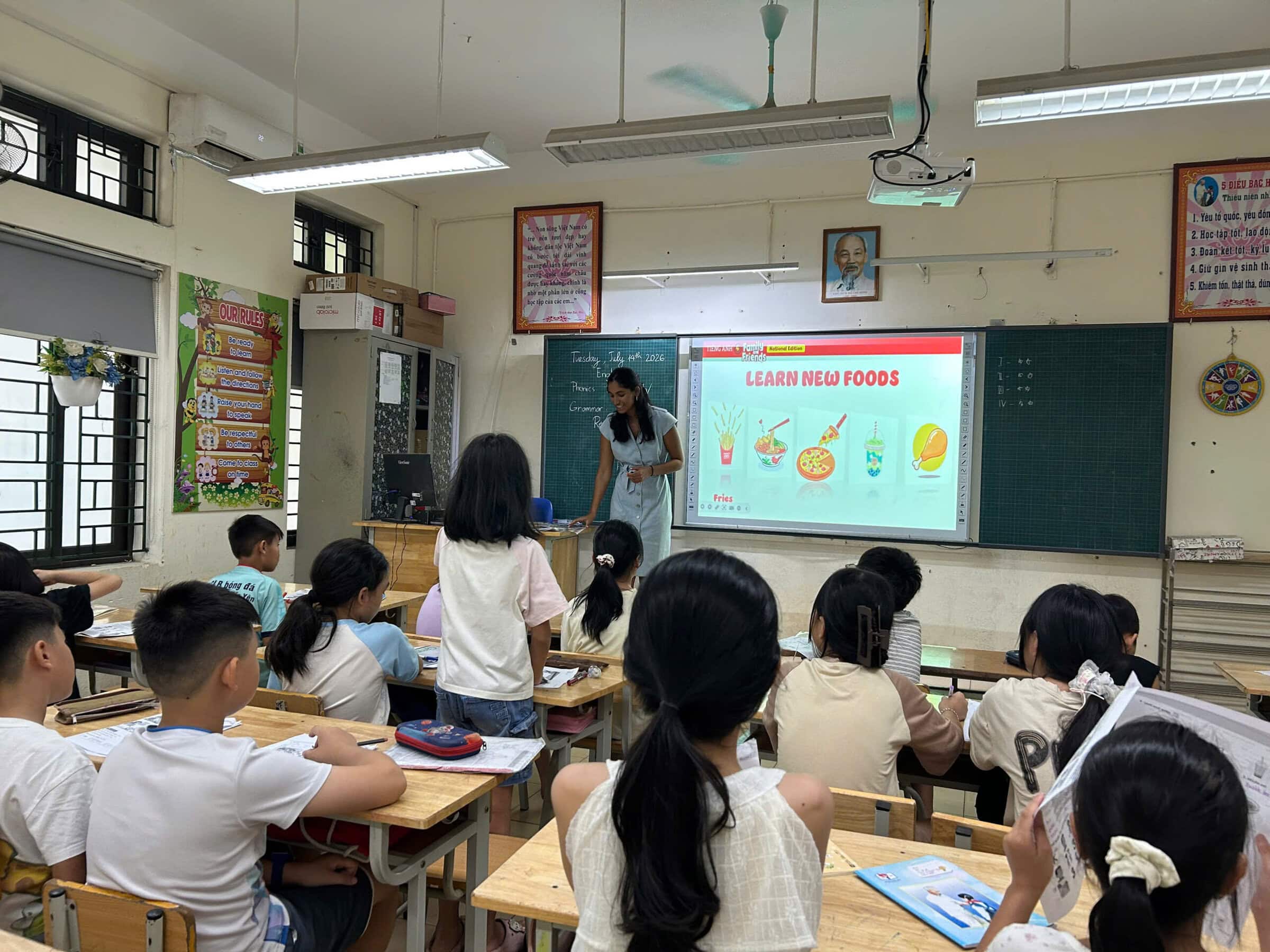 A young woman stands at the front of a classroom beside a screen reading “Learn New Foods,” with pictures of fries, noodles, pizza and bubble tea, as rows of elementary students watch from their desks.