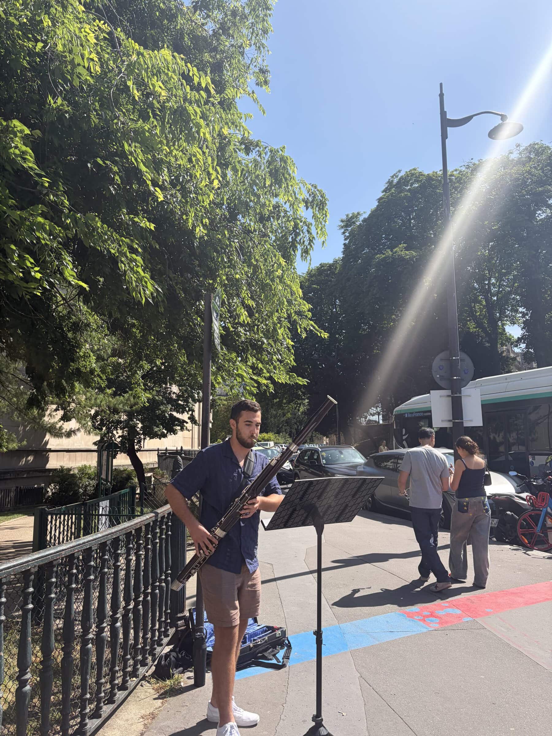 A young man plays bassoon at a music stand on a sunny, tree-lined Paris sidewalk as pedestrians walk past.