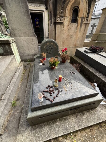 A plain dark granite grave engraved “Claude Debussy,” covered with potted flowers, candles, pebbles and chestnuts in a heart shape, tucked between larger stone mausoleums.