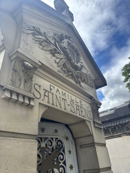 The stone facade of a crypt engraved “Famille Saint-Saëns,” topped with a carved lyre and laurel branches, above an iron door decorated with stars.