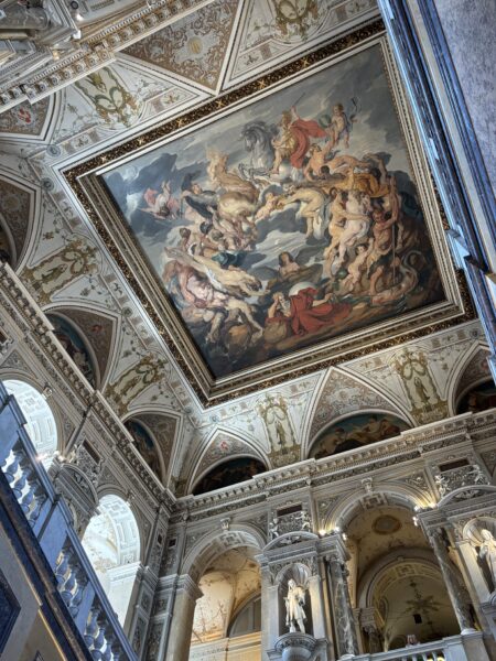 Looking up at an ornate museum ceiling with a large framed painting of swirling mythological figures and a white horse, surrounded by gilded arches and statues.