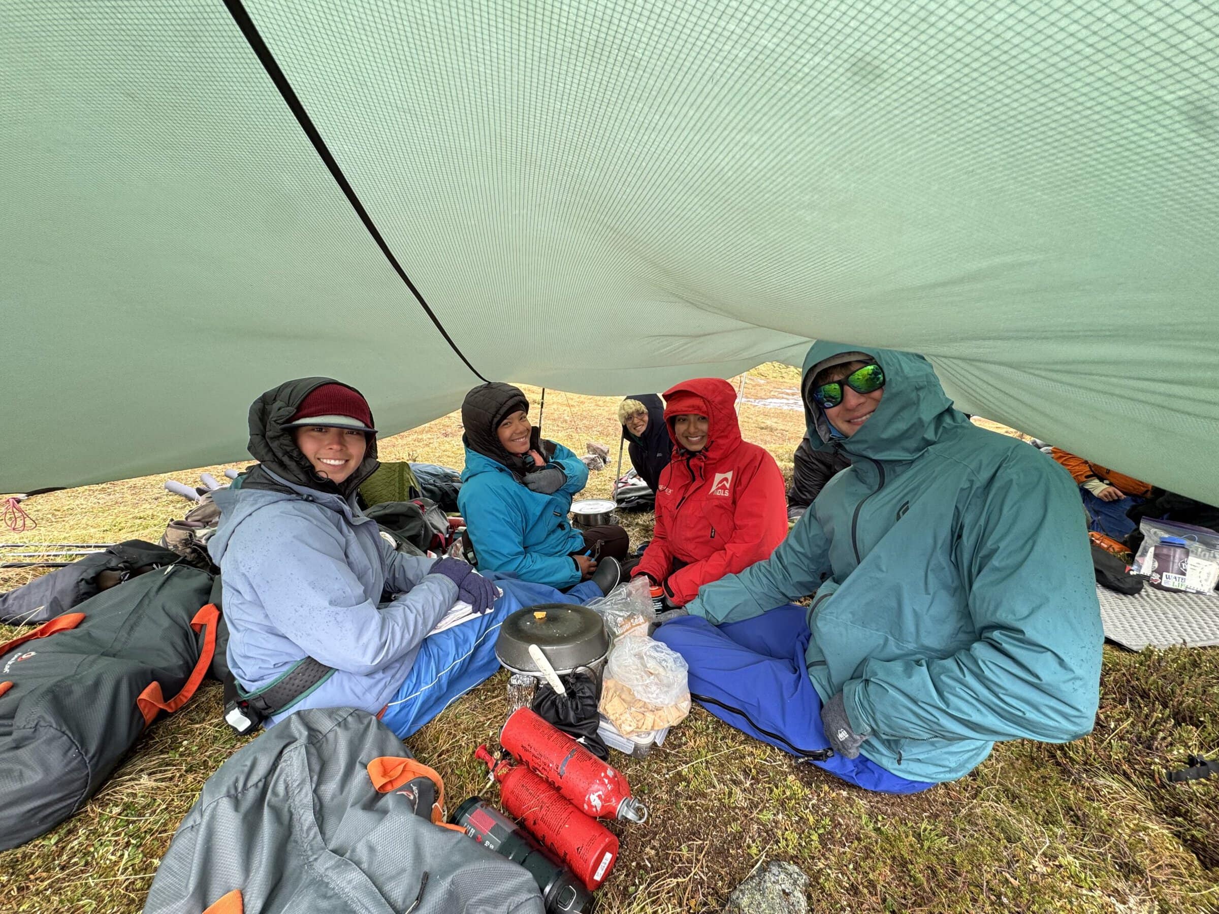 Five people in rain jackets and hoods sit on the tundra under a green tarp, smiling around a camp stove, pots and bags of crackers.