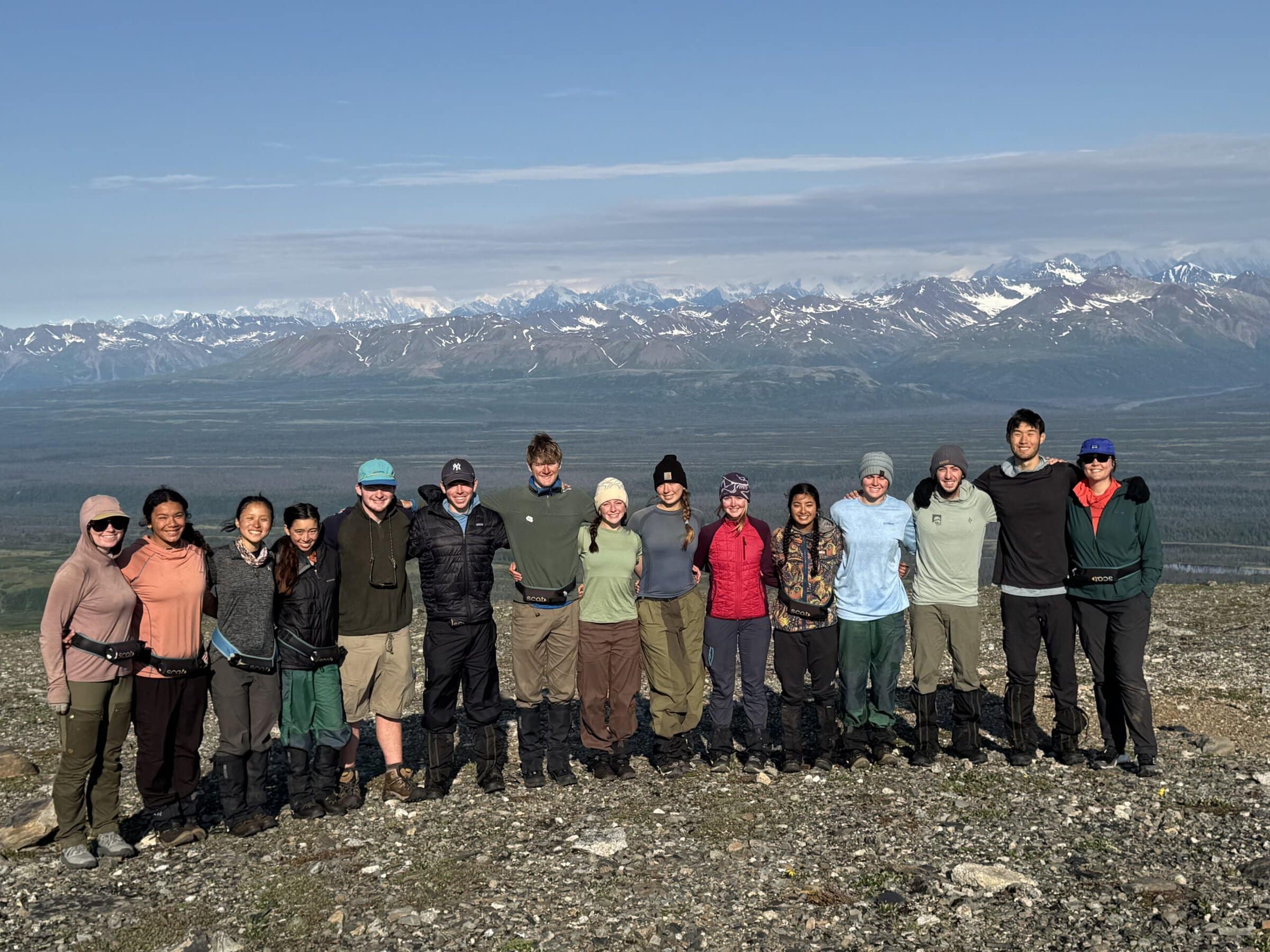A group of hikers stands arm in arm on a rocky ridge, with a wide valley and snow-capped mountains behind them.