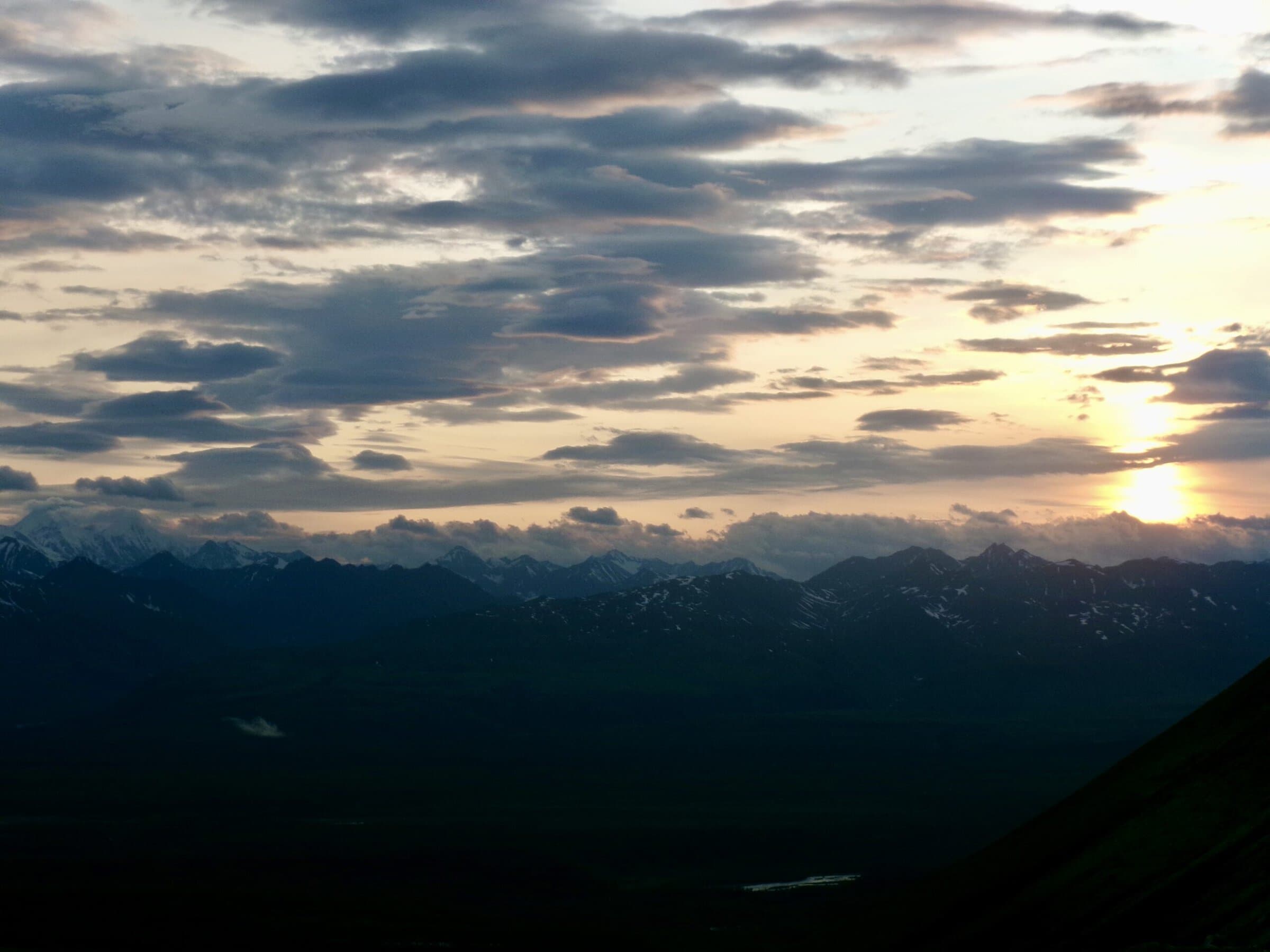 The sun sits low behind layered clouds above a jagged, snow-streaked mountain range and a dark valley.