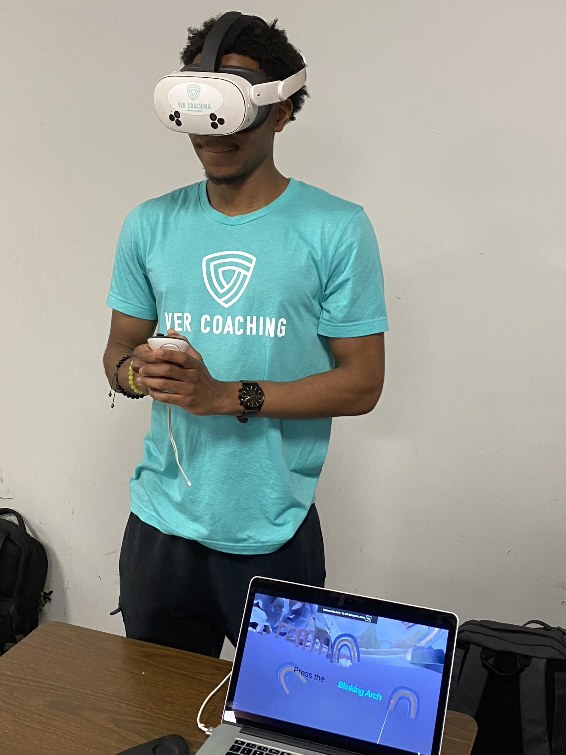 A young man in a teal Ver Coaching T-shirt wears a white VR headset and holds a controller while a laptop in front of him displays a drill reading “Press the Blinking Arch.”