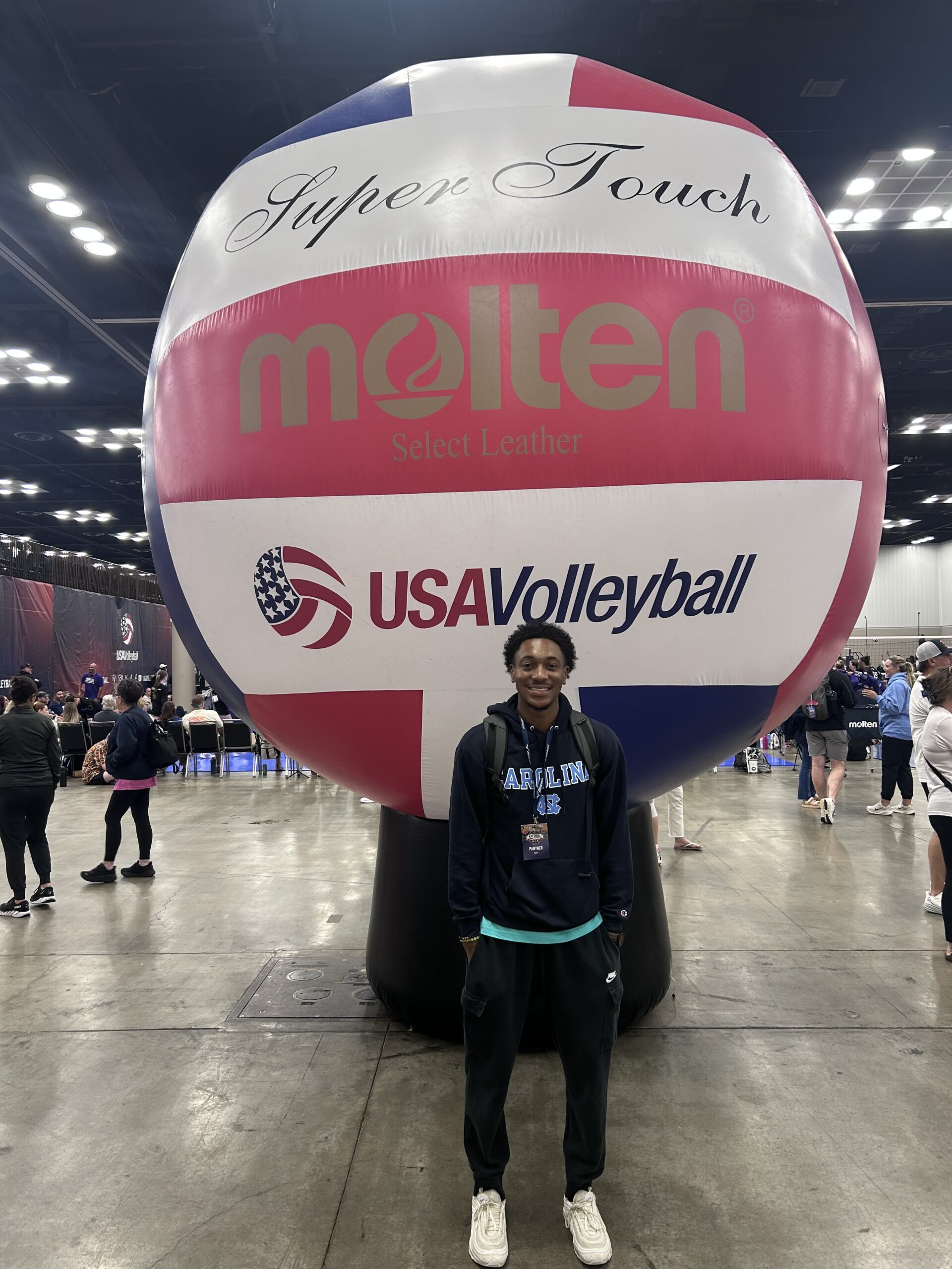 A smiling young man in a navy Carolina hoodie and event credential stands in front of a giant inflatable volleyball printed with USA Volleyball and Molten logos inside a busy convention hall.