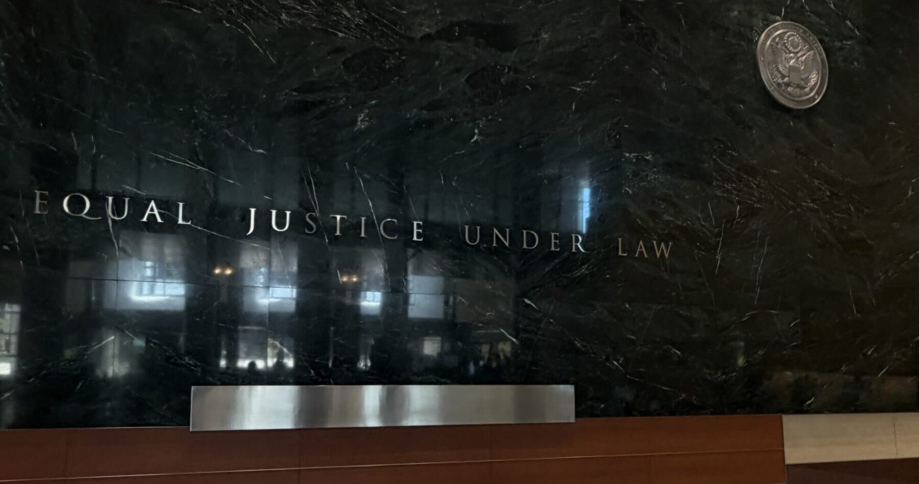 The words “Equal Justice Under Law” in silver letters across a polished black marble wall, with a federal seal to the right, in a courthouse lobby.